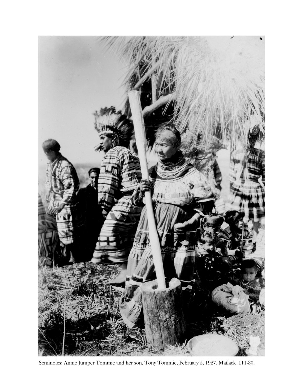 Annie Jumper Tommie使用砂浆和pestel制作玉米面，她的儿子Tony Tommie戴着鹰羽毛帽，1927年2月5日 作者 克劳德·C·马特勒克