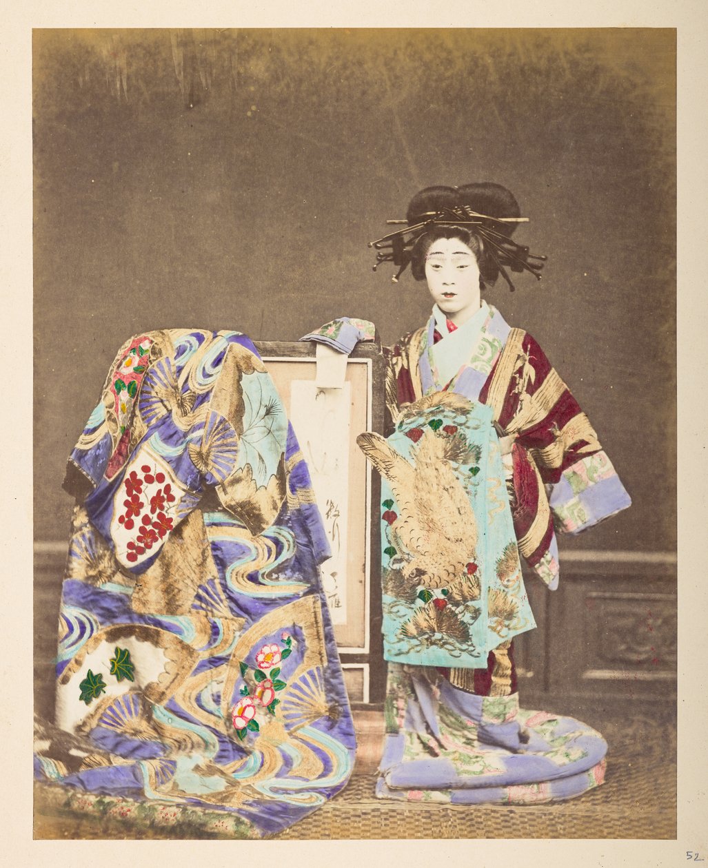 身着传统服装的日本妇女， ] 
一位身着精致服装和发型的女子的全身站立摄影棚肖像，她身旁的画框上还披着另一件长袍，摄影师：卢卡斯收藏：日本传统服饰的摄影记录 作者 英国摄影师