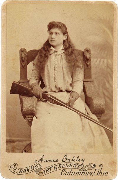 安妮-奥克利 : Annie Oakley:.这位传奇的神枪手端庄地坐在一张文艺复兴风格的大椅子上，膝上放着一把猎枪。她精致的女性特征和飘逸的秀发掩盖了她粗犷的户外形象 ©Buyenlarge/UIG/Leemage 作者 American Photographer