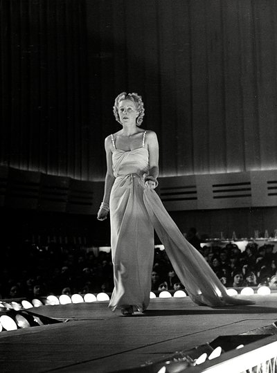 Model photographed on a runway in evening dress with rhinestone shoulder straps; the article is a creation of the couture house "Fumach". (b/w photo) 作者 路易斯·波吉诺（Luis Bogino）